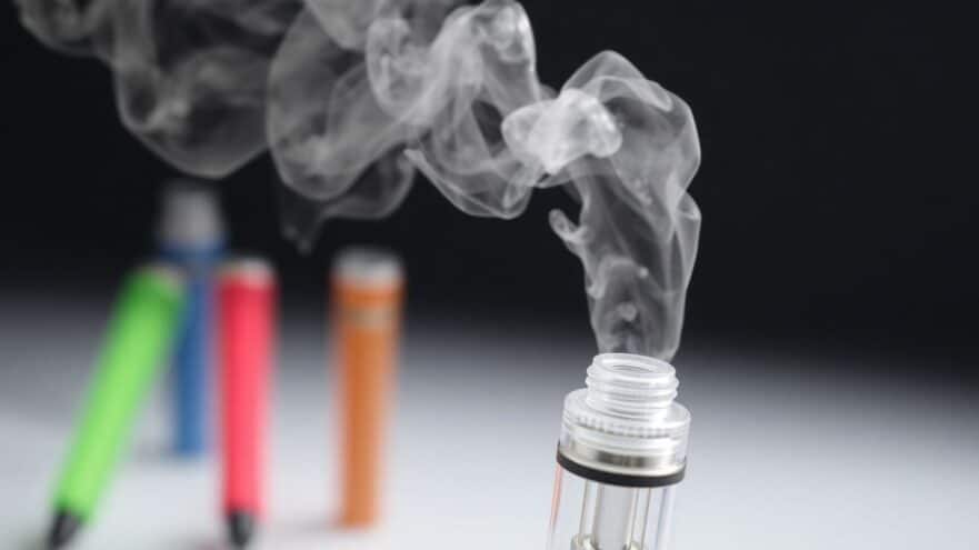 Vape pen with vapor, colorful vapes in background.